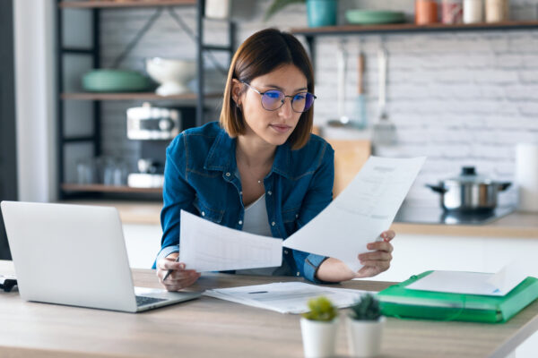 Business woman working with computer while consulting some invoices and documents in the kitchen at home