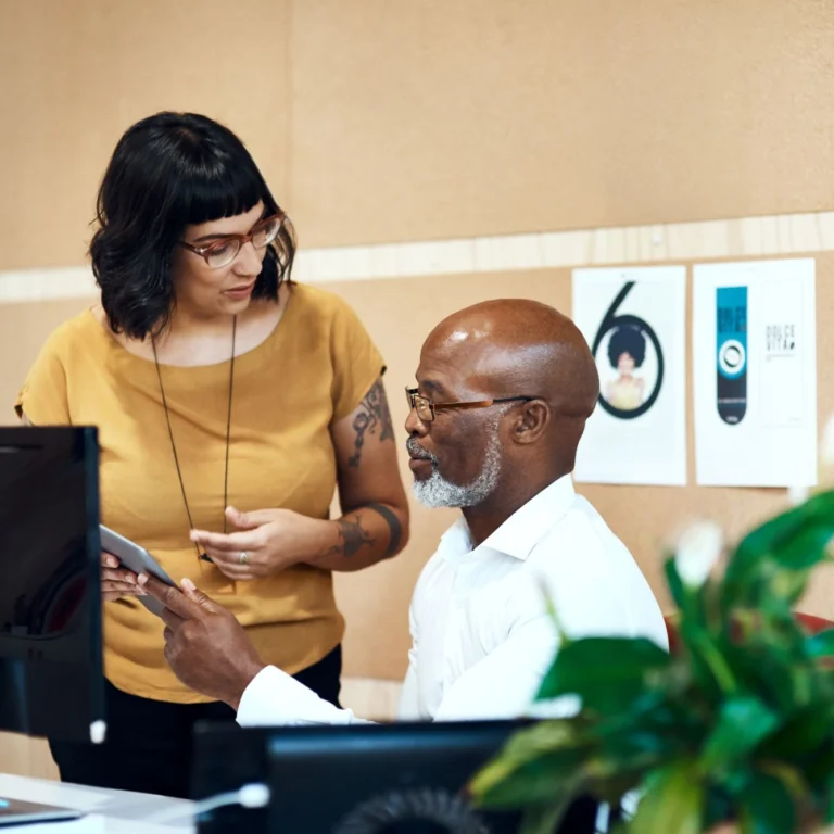 A man and a woman stand next to a computer, looking at a tablet together.