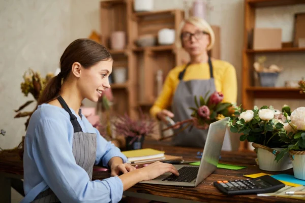 A young woman in a blue shirt and grey apron uses a laptop and calculator in a floristry shop.