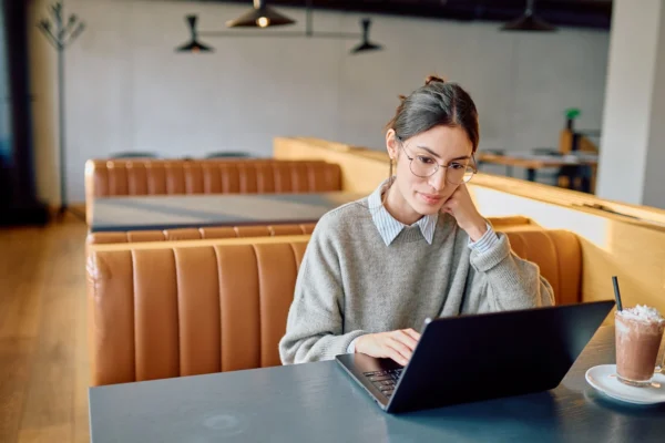 A woman wearing glasses and a sweater sits in a cafe booth, focused on her laptop. 